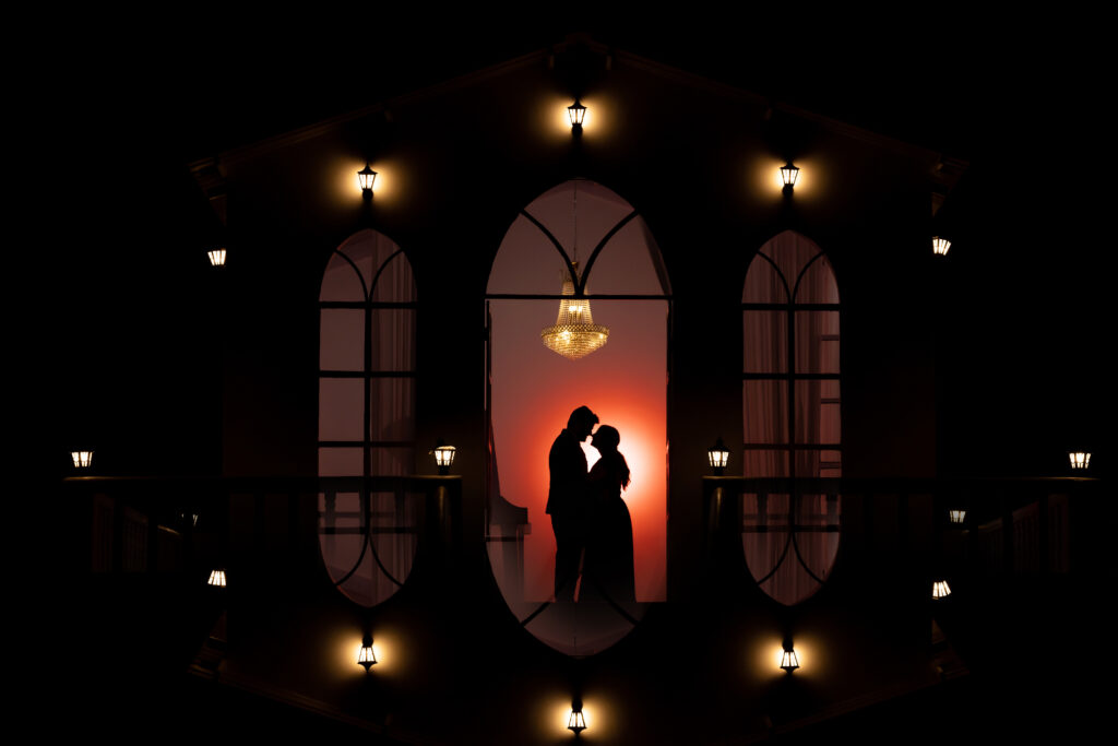 A dramatic silhouette of a couple embracing during a pre-wedding shoot in Jaipur. They are framed by three arched windows of a dark building with small glowing lamps, a crystal chandelier above them, and a warm orange light glowing behind them.