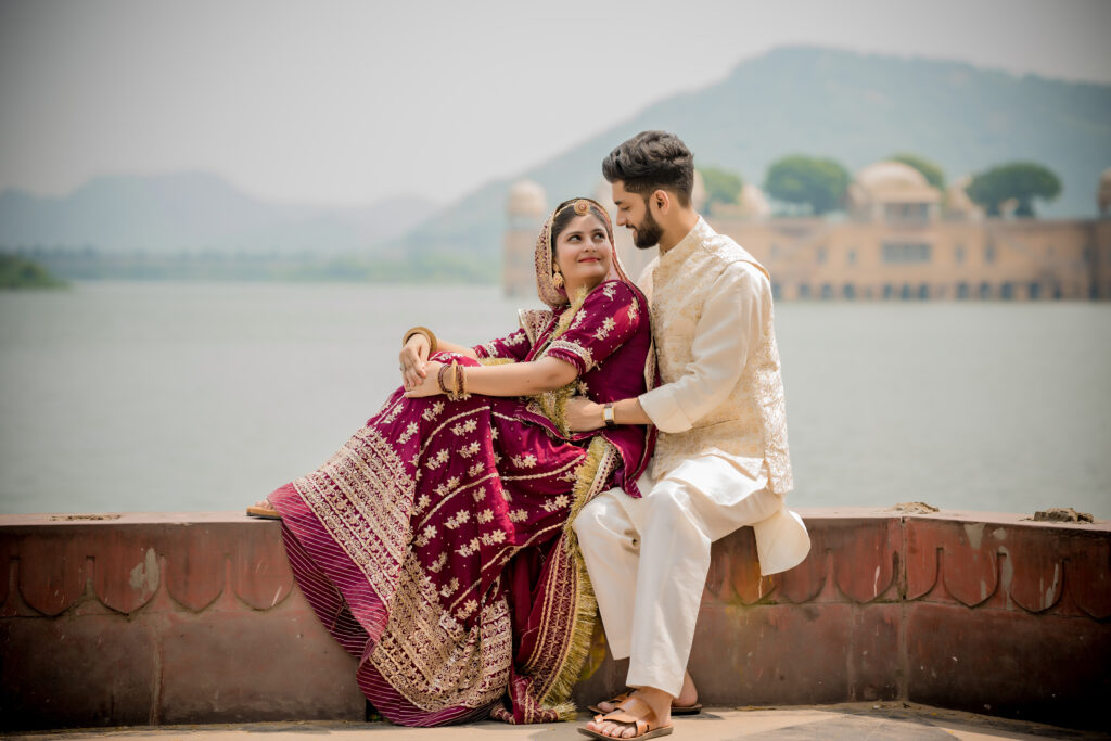 A couple sitting by Man Sagar Lake during a pre-wedding shoot in Jaipur. The bride is in a maroon and gold lehenga, and the groom is in a cream sherwani, with the Jal Mahal palace visible in the background.