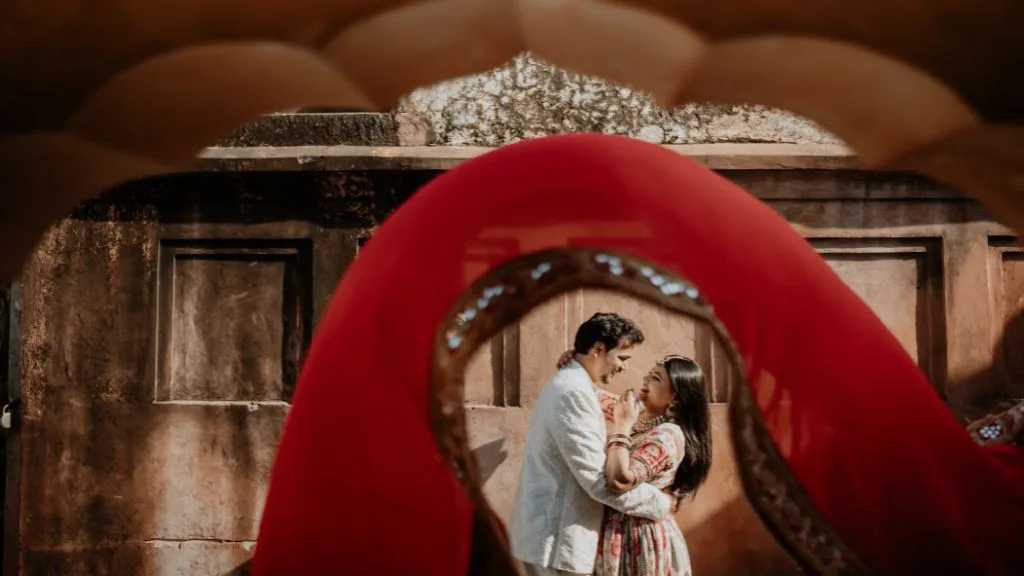 A creative Amer Fort photoshoot featuring a couple framed through a vibrant red fabric veil. The man wears a white sherwani and the woman a traditional patterned dress as they share a romantic moment against the historic stone walls of Jaipur.