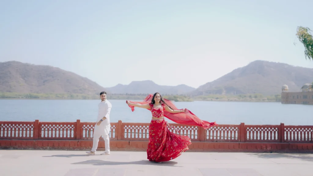 A bride in a red lehenga twirling her dupatta while the groom in white walks beside her during a Jal Mahal pre wedding shoot in Jaipur.
