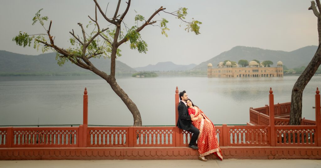 A couple leaning against a red railing during their Jal Mahal pre wedding shoot, with a leaning tree and the water palace in the background.